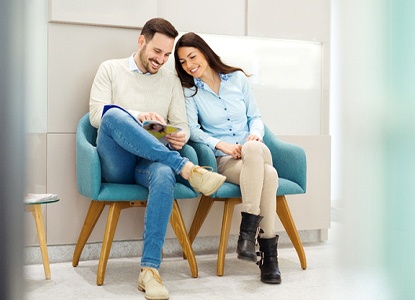 A man and woman sit in a dentist’s office lobby while reading a magazine in preparation for their initial consultation with the dentist