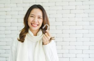 Woman standing in front of a white brick wall wearing a white sweater and smiling as she holds an Invisalign tray